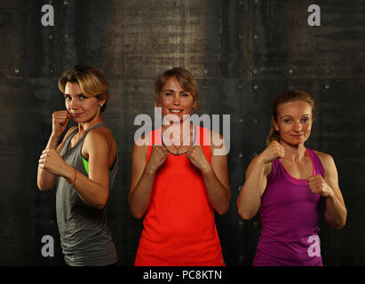 Close up ritratto frontale di tre giovani e di mezza età le donne atletica in abbigliamento sportivo in palestra su sfondo scuro, in piedi in atteggiamento di inscatolamento guardando c Foto Stock