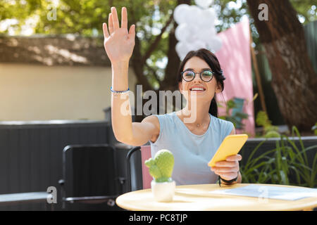 Chiamata cameriere. Piacevole bella donna che indossa gli occhiali sollevando la sua mano durante la chiamata cameriere Foto Stock