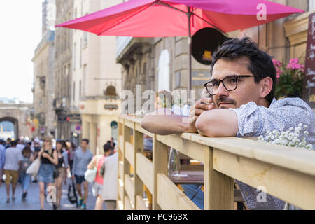 Saint Malo, Francia - Luglio 15th, 2018: un francese di giovane uomo che indossa gli occhiali e il fumo con le braccia su una ringhiera in legno guardando la gente seduta Foto Stock