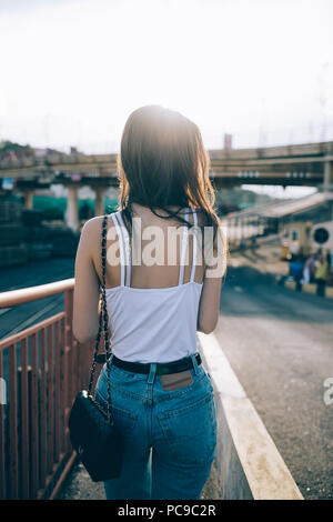 Vista posteriore snello giovane donna con lunghi capelli scuri a piedi giù per la strada al tramonto. Ragazza adolescente jeans blu e bianco superiore vagare in città Foto Stock