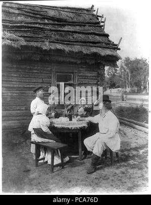 In scena il genere view include l uomo con la balalaika, samovar sul tavolo, ragazzo versando bere, uomo del peering attraverso la finestra e gli altri seduti a tavola al di fuori di un tetto di paglia casa log. Contadina russa s 1875 Foto Stock