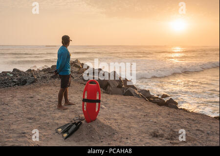Bagnino di turno visto da dietro, guardando oltre l'oceano da un sunset beach. Galleggiante rosso e nero di pinne sulla sabbia, onde si infrangono nelle rocce. Foto Stock