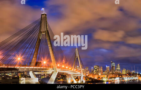 Moderno ponte urbano nella città di Sydney CBD contro le torri distanti e nuvoloso susnet sky con l'australiano anzac bandiera su alta colonna con luci e illumina Foto Stock