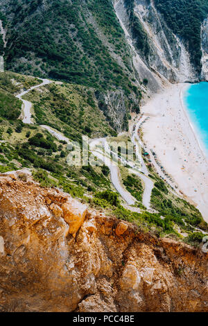 Serpentina strada a zigzag giù per la famosa spiaggia di Myrtos. Rocce di colore arancione burrone sul lato. Sunny beach dal punto di vista platform, Cefalonia, Grecia. Foto Stock