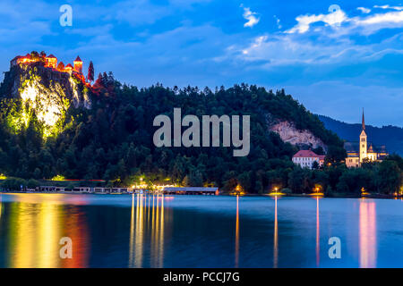 Il castello di Bled illuminato durante l ora di blu che si affaccia sul lago in un giorno di estate. Castello sloveno su una scogliera illuminata di notte con acqua riflessioni Foto Stock