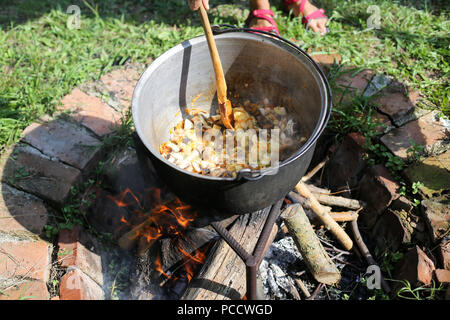 Donna agitazione in una ghisa calderone dove uno stufato di gulasch è in ebollizione, oltre il caminetto aperto nel cortile di una casa rurale in Romania Foto Stock