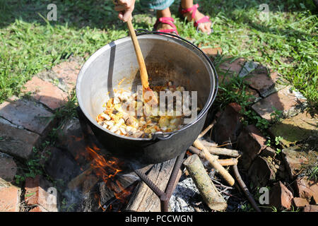 Donna agitazione in una ghisa calderone dove uno stufato di gulasch è in ebollizione, oltre il caminetto aperto nel cortile di una casa rurale in Romania Foto Stock