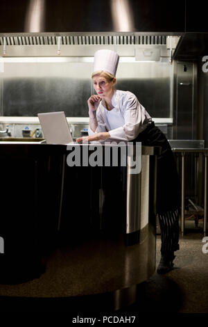 Una femmina caucasica chef lavorando su un computer portatile dopo ore in una cucina commerciale. Foto Stock