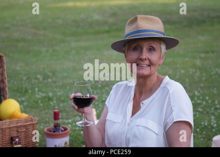 Con un cappello per il sole sul suo capo e un bicchiere di vino rosso, la donna graziosa gode di un picnic. Foto Stock