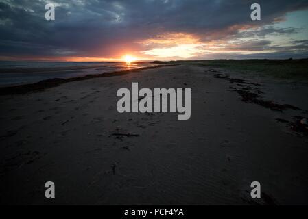 Una vista guardando verso il basso di una spiaggia di sabbia con regolazione del sole. Il sole tramonta su una lunga spiaggia di sabbia con alghe e impronte sulla sabbia. Foto Stock