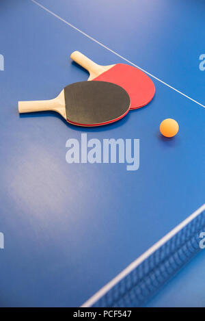 Sfera arancione per il tennis da tavolo e due racchette in legno di colore rosso e nero su un tavolo blu con una griglia Foto Stock