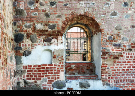 Una finestra con un arco e il reticolo di ferro in una spessa, vecchia fortezza di mattoni parete attraverso la quale il cortile è visibile. Foto Stock