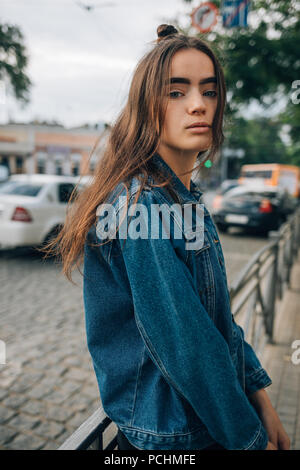 Bella ragazza in un denim blu con camicia in posa nella città vicino alla strada. Moda giovane donna con lunghi capelli scuri guardando la telecamera appoggiata su parap Foto Stock