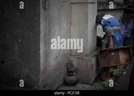 Vecchio senzatetto asiatica donna in piedi oltre un angolo, guardando verso il basso, indossando un blu e abito grigio, tenendo in mano un vassoio in legno Grande muro grigio sulla sinistra. Foto Stock