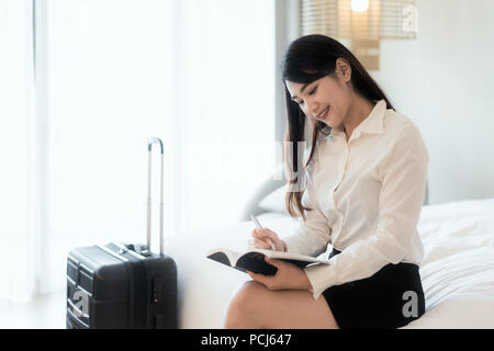 Close up di un felice bella Asian business donna sorridente allegramente in appoggio al suo hotel suite durante il viaggio di affari rendendo note e pensando strate Foto Stock