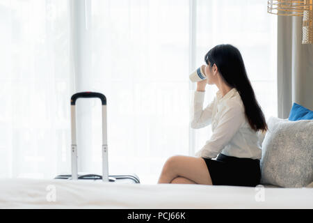 Rilassarsi dopo una dura giornata di lavoro. Bella asiatica giovane imprenditrice sorridente in tuta di bere il caffè e guardare lontano mentre è seduto sul divano in hotel r Foto Stock