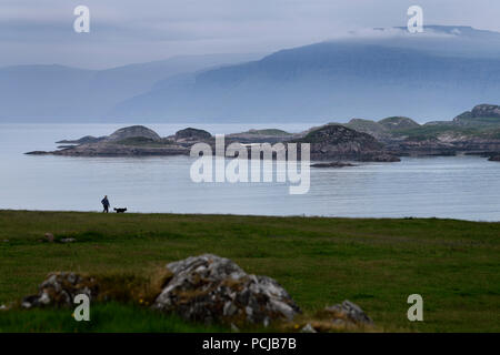 Donna che cammina il suo cane con vista di Isle of Mull scogliere e ben più montagna di oltre il suono di Iona dall isola di Iona Abbey al tramonto Scozia UK Foto Stock