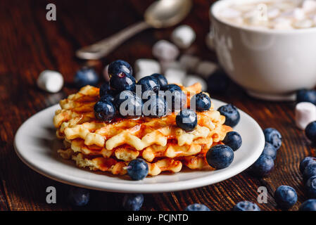 Waffles fatti in casa sulla piastra con mirtilli freschi e sciroppo. Tazza di cioccolata calda e alcuni frutti di bosco con Marshmallow su sfondo. Foto Stock