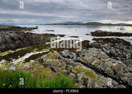 Spiaggia di sabbia e costa rocciosa sotto le nuvole sull isola di Iona con barche sul suono di Iona e Fionnphort Isle of Mull montagne Scotland Regno Unito Foto Stock