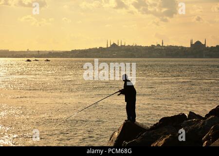 L'uomo è la pesca sulle rocce vicino al faro e la silhouette della città in background Foto Stock