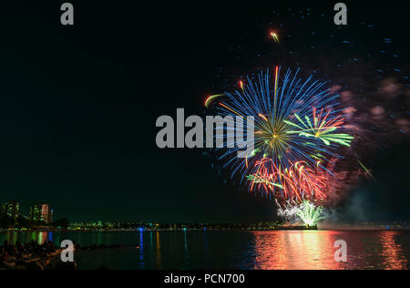 Multicolore di fuochi d'artificio sull'oceano nella grande città, sagome di gente sulla spiaggia di moderni edifici sullo sfondo, incandescente e riflettendo Foto Stock