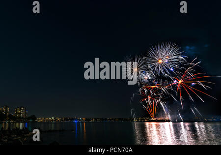 Multicolore di fuochi d'artificio sull'oceano nella grande città, sagome di gente sulla spiaggia di moderni edifici sullo sfondo, incandescente e riflettendo Foto Stock