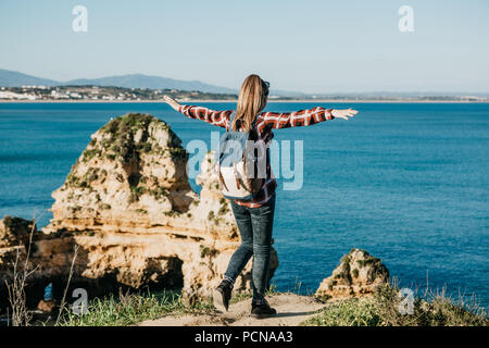 Una ragazza di turista o viaggiatore con lo zaino si ammira la splendida vista dell'Oceano Atlantico in Portogallo e solleva le mani in alto mostra come le piace e come essa è libera. Foto Stock