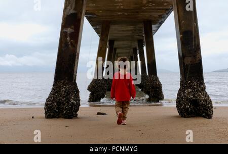 Un giovane bambino in piedi sotto un molo vicino all'oceano, la Cardwell Jetty, Cardwell, Queensland, Australia Foto Stock