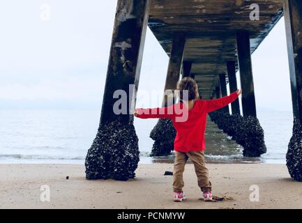 Un giovane bambino in piedi sotto un molo vicino all'oceano, la Cardwell Jetty, Cardwell, Queensland, Australia Foto Stock