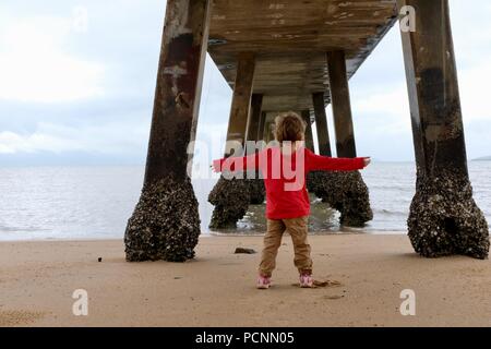 Un giovane bambino in piedi sotto un molo vicino all'oceano, la Cardwell Jetty, Cardwell, Queensland, Australia Foto Stock