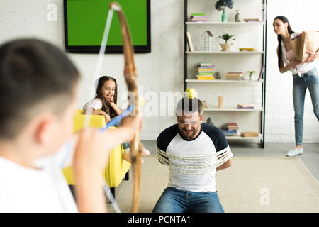 Bambini che giocano con legato padre e fingere di tiro con arco giocattolo in salotto Foto Stock