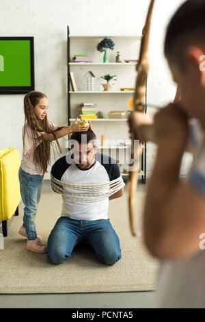 Bambini disobbedienti giocando con legato padre e fingere di tiro con arco giocattolo a casa Foto Stock