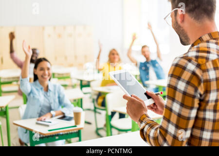 Un gruppo di giovani studenti alzando le mani per rispondere sulla questione degli insegnanti mentre lui in piedi con la compressa in primo piano Foto Stock