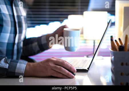 Lavorare fino a tarda notte e di bere il caffè. Uomo che utilizza il computer portatile e la coppa di ritegno o mug in mano in casa o in ufficio. Foto Stock