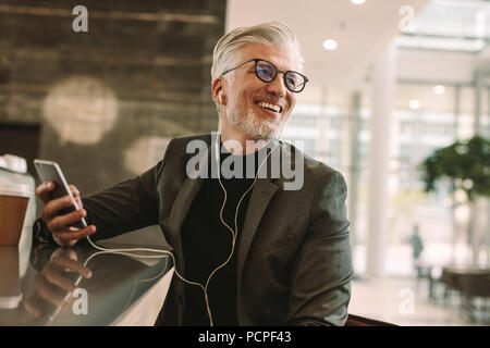 Ritratto di sorridente imprenditore maturo con gli auricolari e il telefono cellulare seduti al coffee shop e guardando lontano. Foto Stock