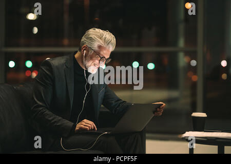 Sorridente imprenditore maturo lavora fino a tardi sul computer portatile in ufficio lobby. Professionista aziendale guardando laptop e sorridente. Foto Stock