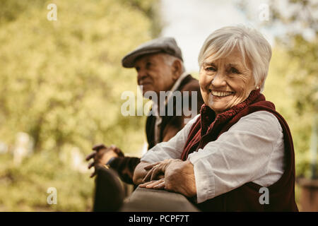 Ritratto di bella donna senior in piedi da una ringhiera in legno con il suo partner sul retro in posizione di parcheggio. Sorridente vecchia donna al parco con il marito. Foto Stock