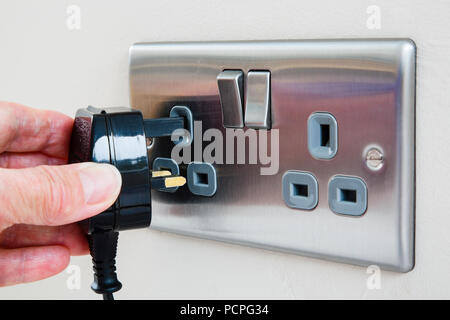 La mano di una persona in possesso di un tre pin connettore e collegarlo a una presa a muro elettrica quando viene accesa. Inghilterra, Regno Unito, Gran Bretagna Foto Stock