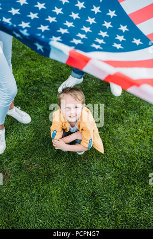 Ritagliato colpo di genitori holding bandiera americana e piccolo figlio accovacciato sull'erba Foto Stock