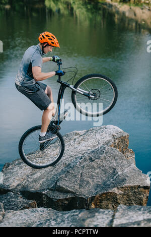 Estrema prova biker bilanciamento sulla ruota posteriore sulla scogliera di roccia sopra il lago Foto Stock