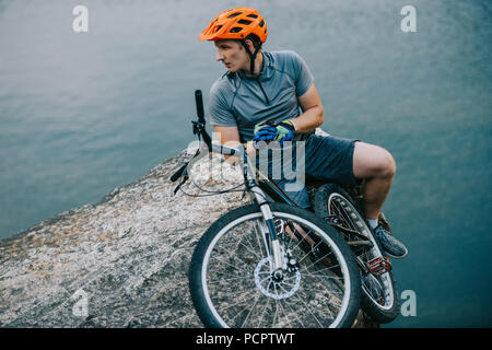 Prova di giovani biker rilassante sulla scogliera rocciosa su acqua e guardando lontano Foto Stock