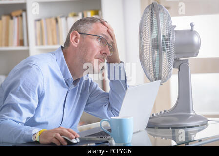 L'uomo soffre dal calore durante il lavoro in ufficio e si tenta di raffreddarsi dalla ventola Foto Stock
