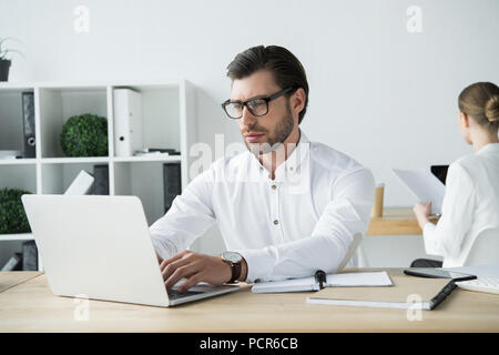 Focalizzato giovane imprenditore lavora con il portatile in ufficio moderno con il collega seduto su sfondo Foto Stock