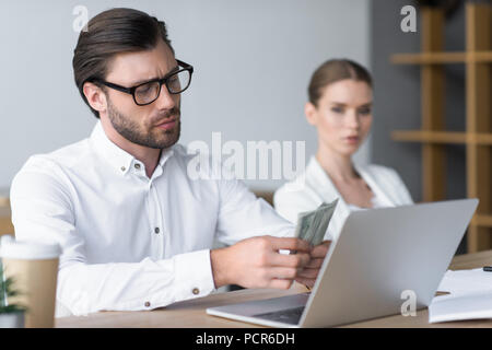Grave giovane impreditore Conteggio contanti mentre il suo collega guardando lui sfocati sullo sfondo Foto Stock