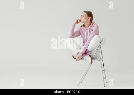 Considerato giovane donna in rosa hoodie seduti su una sedia su bianco e guardando lontano Foto Stock