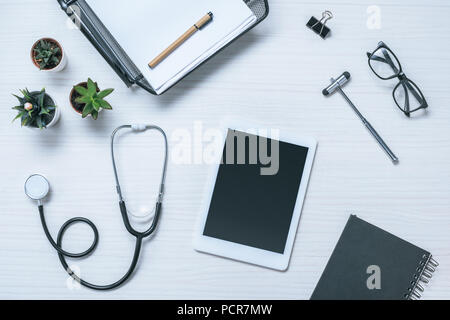 Vista in elevazione del medico sul posto di lavoro con il libro di testo, lo stetoscopio e il martello reflex e digitale compressa sul tavolo Foto Stock