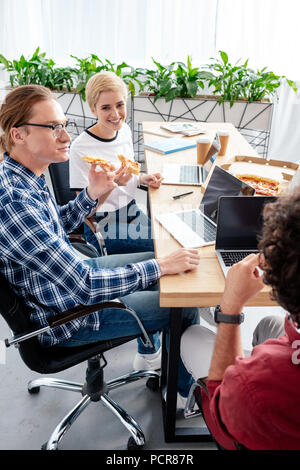 Sorridente giovani colleghi mangiare la pizza seduti insieme al lavoro Foto Stock