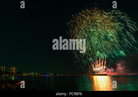 Multicolore di fuochi d'artificio sull'oceano nella grande città, sagome di gente sulla spiaggia di moderni edifici sullo sfondo, incandescente e riflettendo Foto Stock