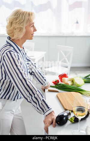 Vista laterale di attraenti i capelli grigi donna appoggiata sul banco di cucina durante la cottura in cucina Foto Stock