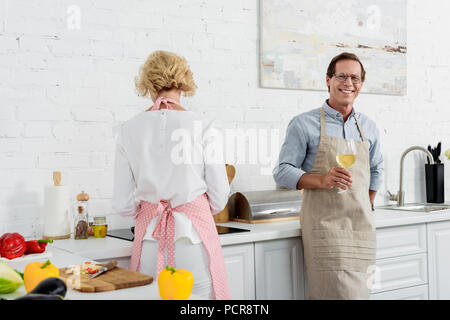 Uomo anziano in grembiule tenendo un bicchiere di vino e sorridente in telecamera durante la cottura con la moglie in cucina Foto Stock